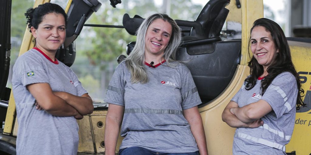 A imagem é uma foto de três mulheres, funcionárias da JSL, uniformizadas com uma camiseta cinza, sorrindo para a câmera. Atrás delas está uma empilhadeira amar